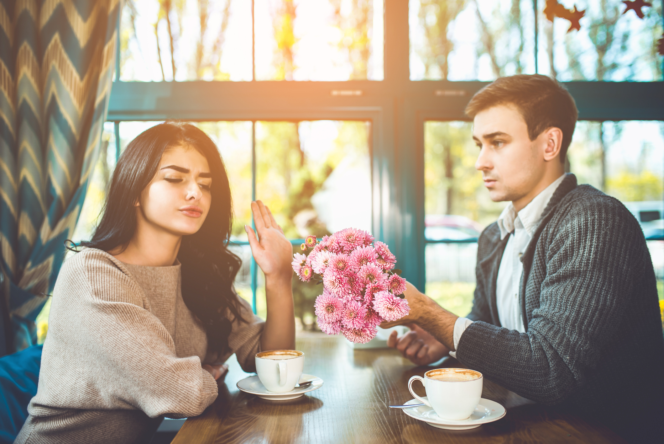 A man and a woman on a date. They are sat opposite each other at a table in what seems to be a coffee shop or restaurant. The man is holding out a bunch of flowers to the woman. The woman does not look impressed and is putting her hand up to the man, suggesting she does not want the flowers.