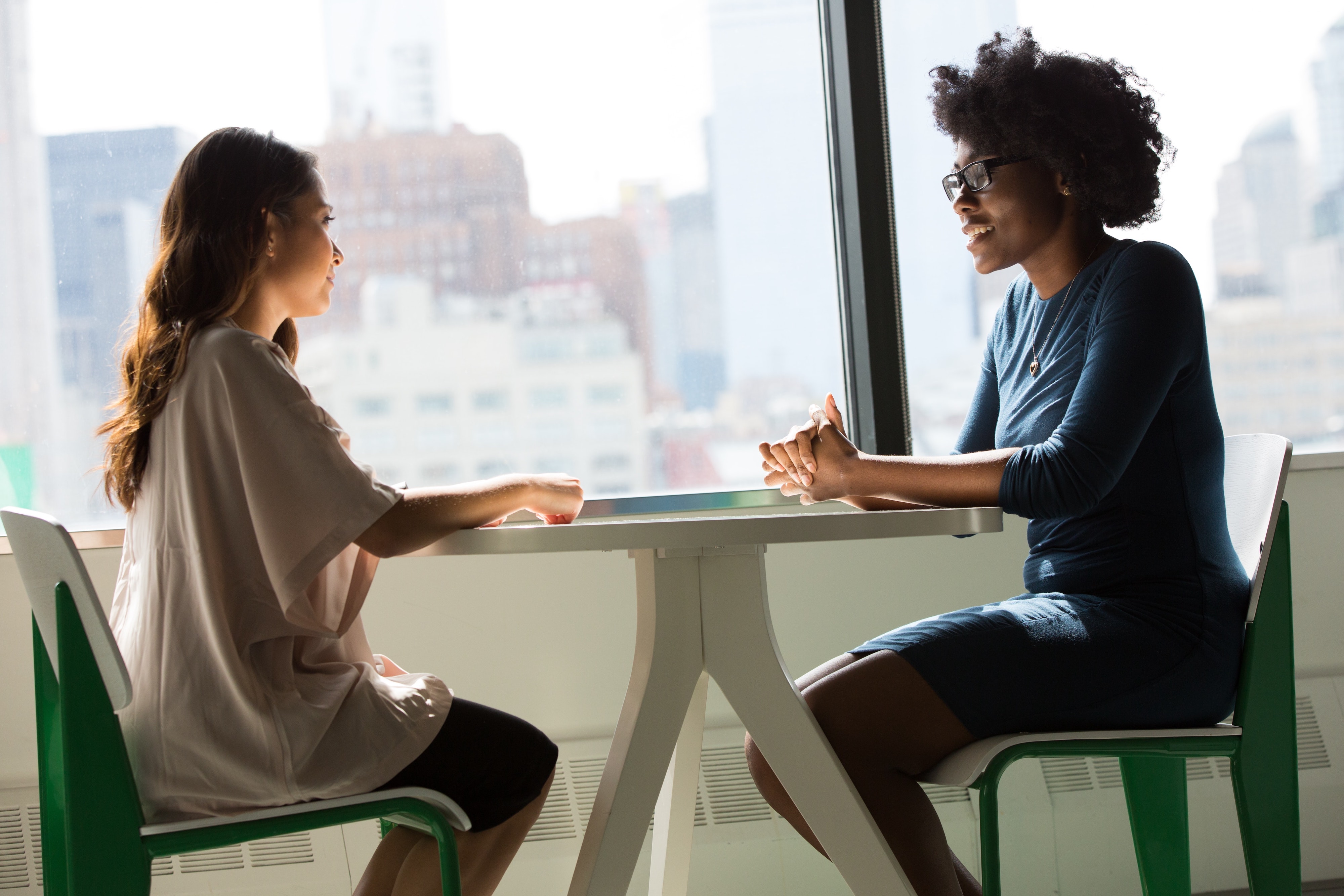 A photo of two women sat at a desk opposite each other. They appear to be in a work environment, and are having an informal conversation.