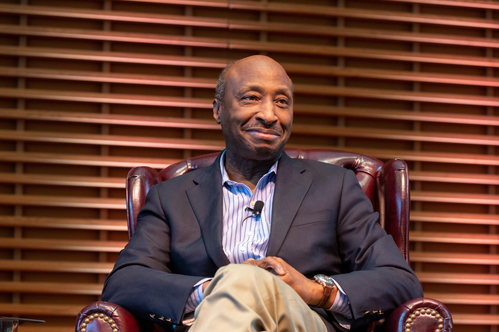 A photo of Kenneth C. Frazier, a successful black man who has worked in law and pharmaceuticals. More recently, he is a vocal advocate for social justice. In the portrait, he is sat in an armchair and is dressed smartly. He is looking away from the camera smiling.