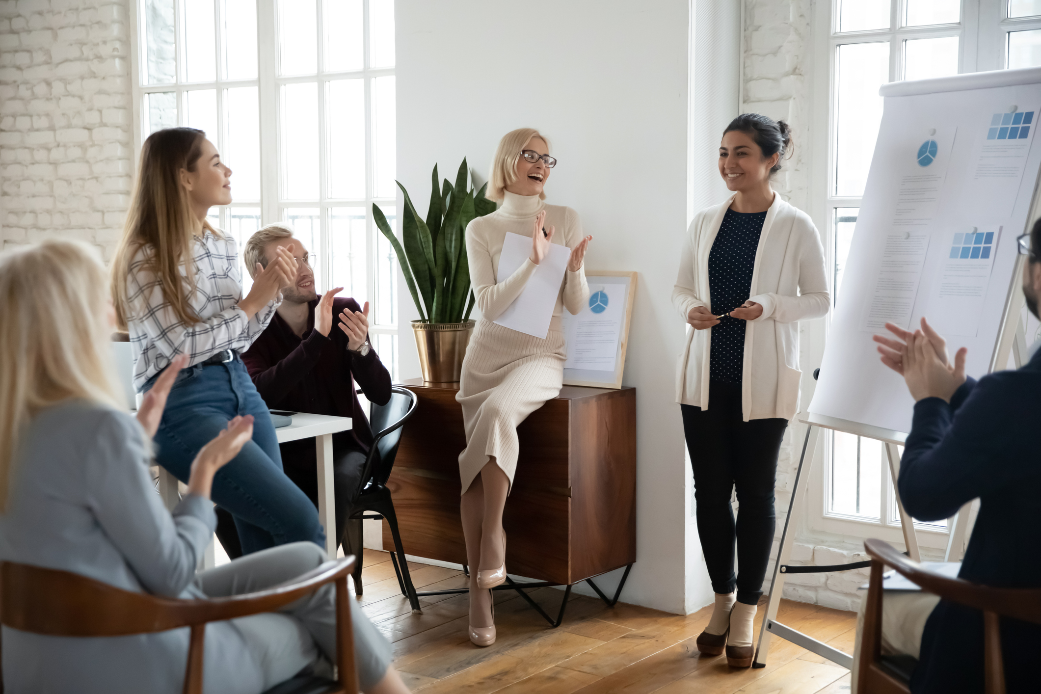 A group of colleagues listening to a member of the team give a presentation. They are clapping and smiling at the colleague, who is stood next to a flipchart presentation.