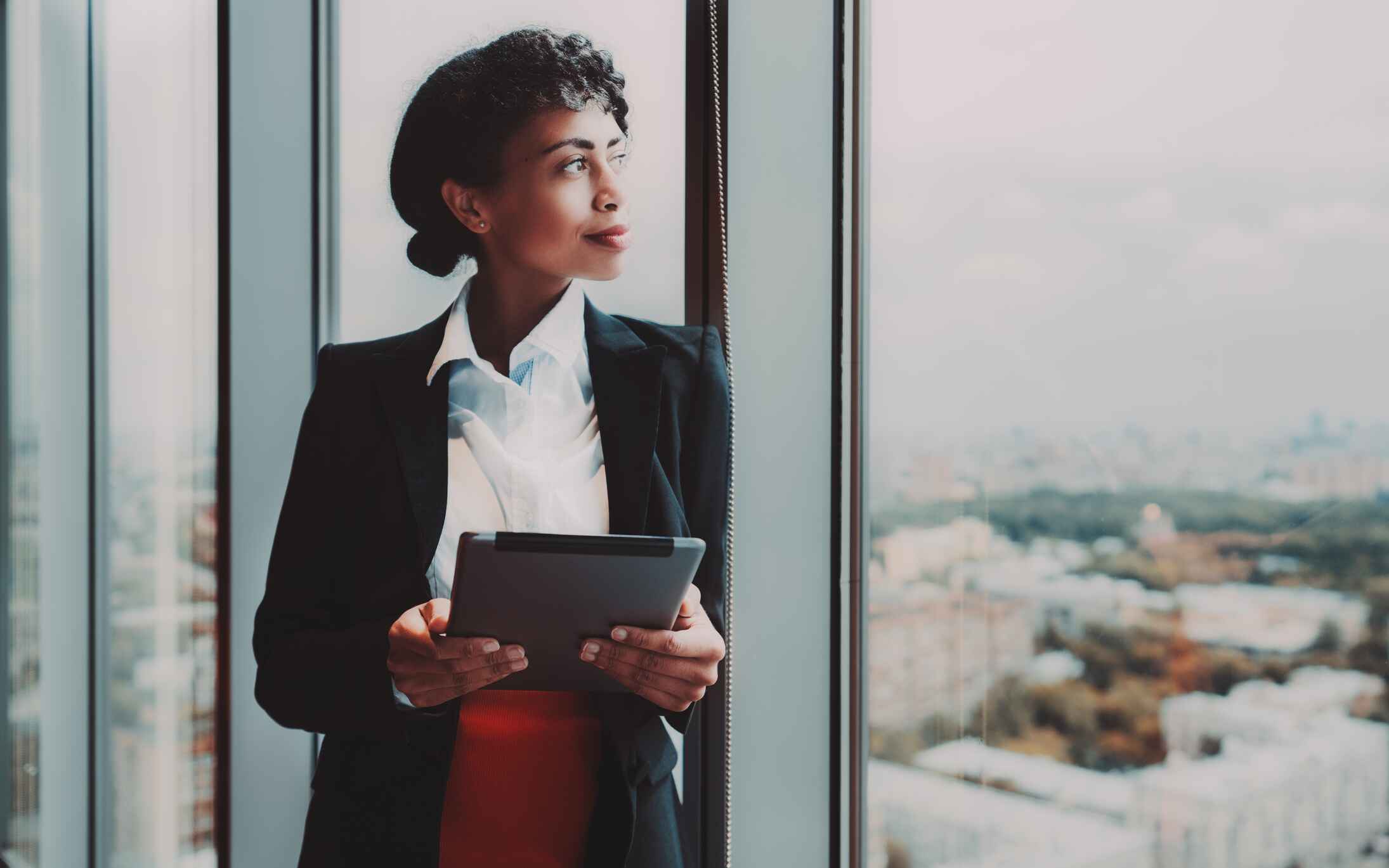 A professional woman dressed in smart wear. She is stood next to a large window looking out at a city skyline. She is holding an electronic tablet, and is smiling to herself in a confident manner while looking out of the window.