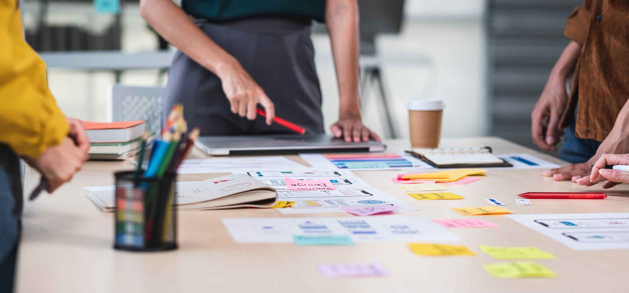 An image of a table with post-it notes, pens and other stationery on it. There are a team of colleagues stood around the table - you can just see their hands and torsos. They are smartly dressed. A female colleague is leading the conversation, and is pointing at some of the work on the table.