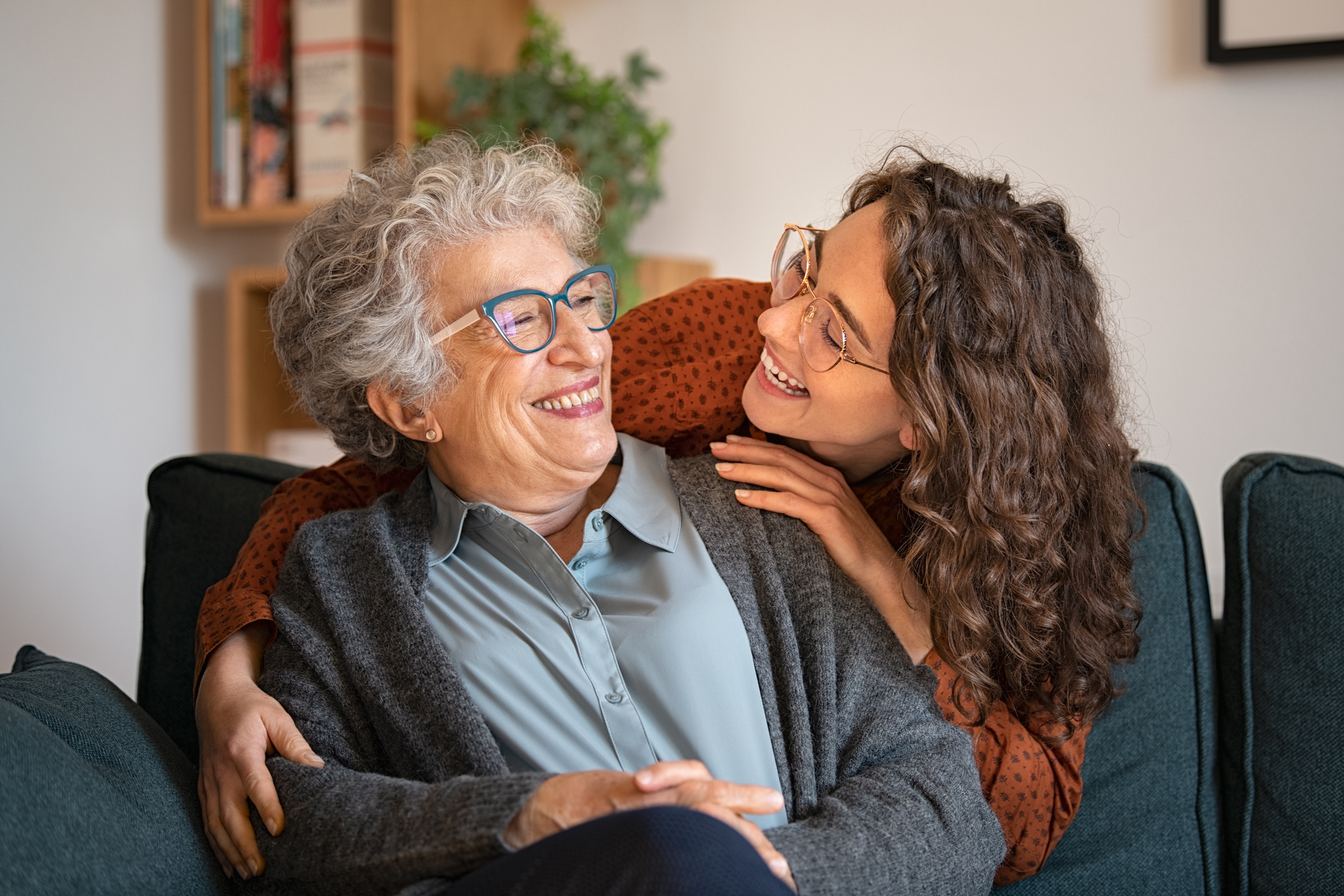 Two women embracing. The older woman is sat in an armchair, while the younger woman is leaning down to put her arms around her. They are smiling at each other. It looks as if they may be related, perhaps mother and daughter.