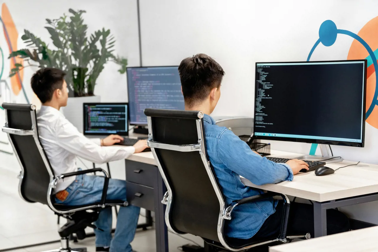 Two men dressed in smart casual clothing sitting on office chairs at a desk. They are both looking at computer screens with several lines of code.