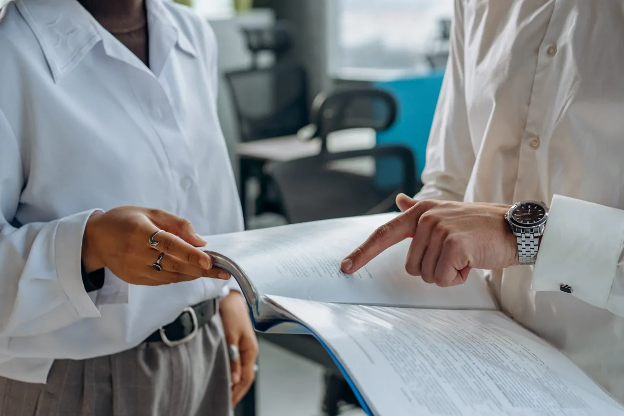 Two people in smart business attire are holding a file containing several documents. The file is open and they are looking at the contents of the double page spread. One person is pointing at something on the page.