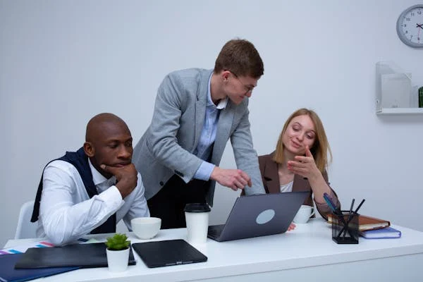 Three colleagues collaborating at a desk in a modern office. One person in a grey blazer points at a laptop screen, explaining something to a female colleague who is smiling. Another male colleague sits thoughtfully, listening, with coffee cups, documents, and a small plant on the desk.