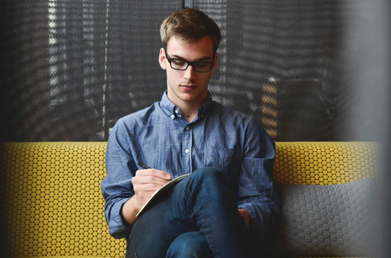 A man is sitting on a yellow bench with a grey cushion beside him. He is dressed in smart-casual clothes and has glasses on. He is balancing an open notepad on top of crossed legs. He is looking down at the page and writing something on it with a black pen.