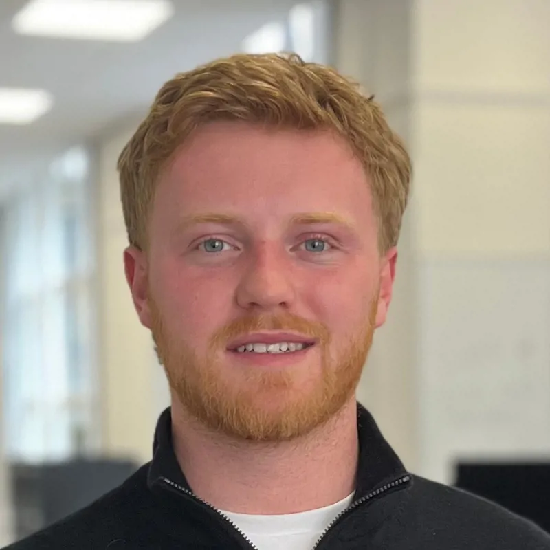 A young man standing in an office smiles at the camera. This is James Hayne, Consultant.