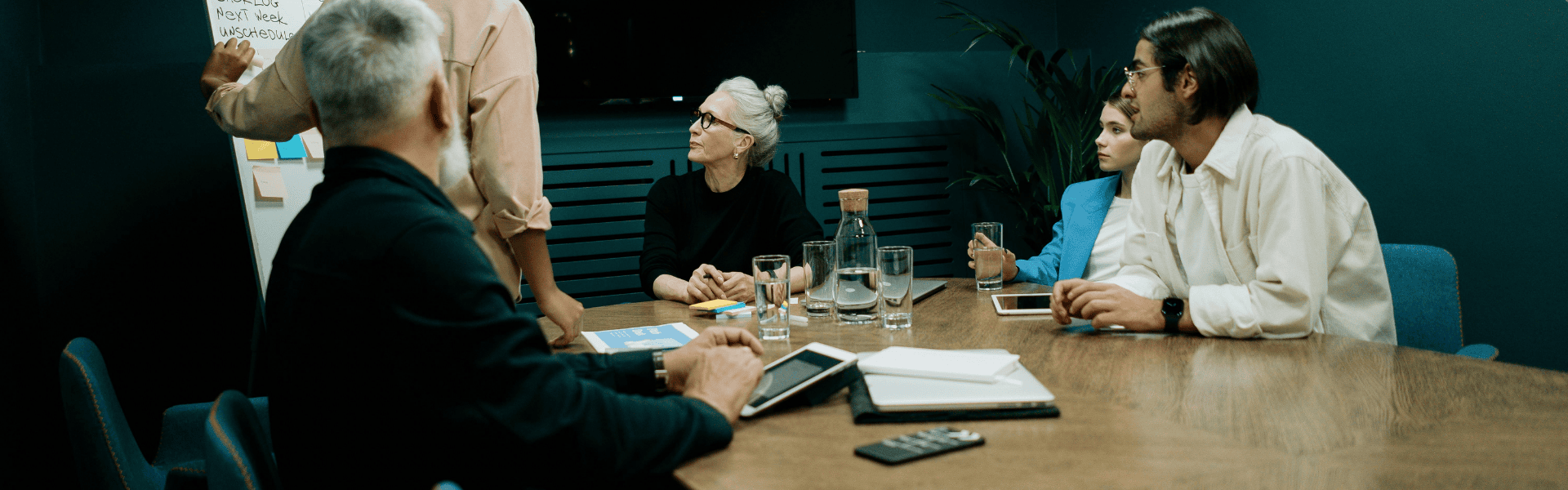 A group of colleagues sit around a desk in a modern office. One person is standing and pointing to something offscreen and the others are all looking in that direction. There is a jug and glasses of water on the table, as well as some electronic tablets and documents.