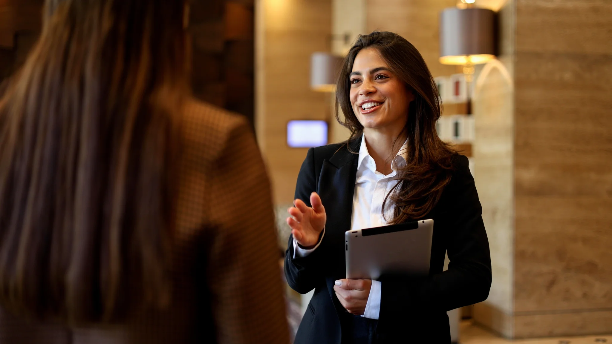 Businesswoman talking with colleague, holding digital tablet in modern office or hotel lobby, smiling during discussion