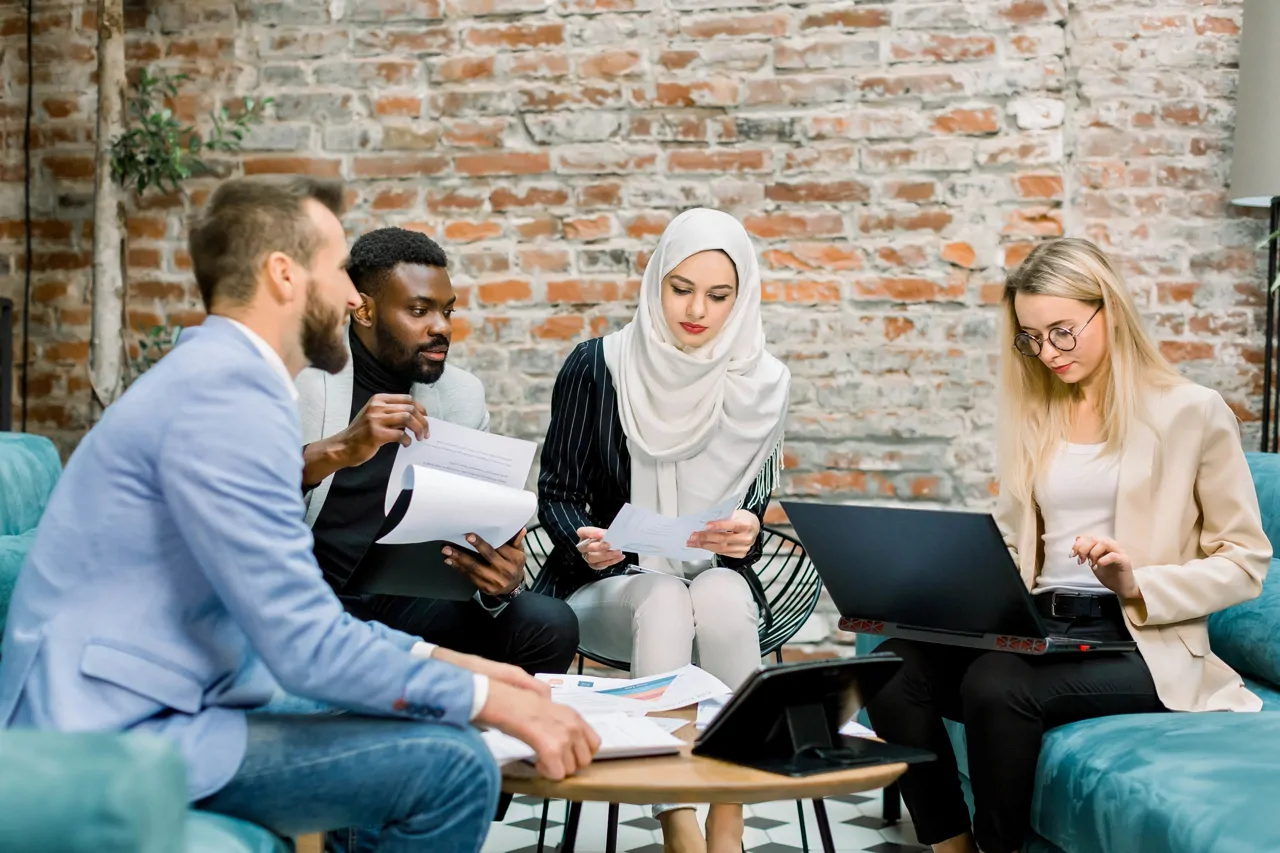 Four people sat on a blue couch at a low table having a meeting with a laptop, notes