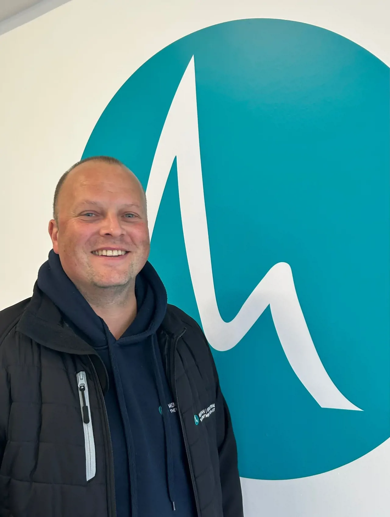 Man smiling wearing Meridian branded jacket stood beside Meridian roundel logo