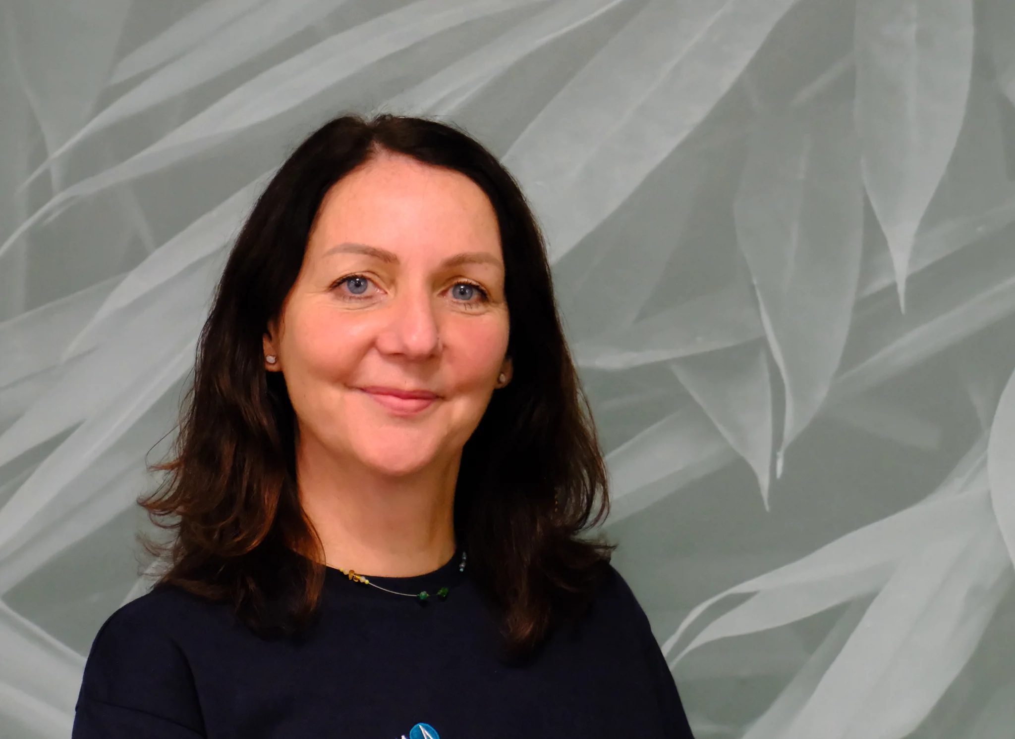 Woman with shoulder-length dark brown hair, wearing a black top and a subtle necklace, smiling gently in front of a grey background with a leafy pattern.