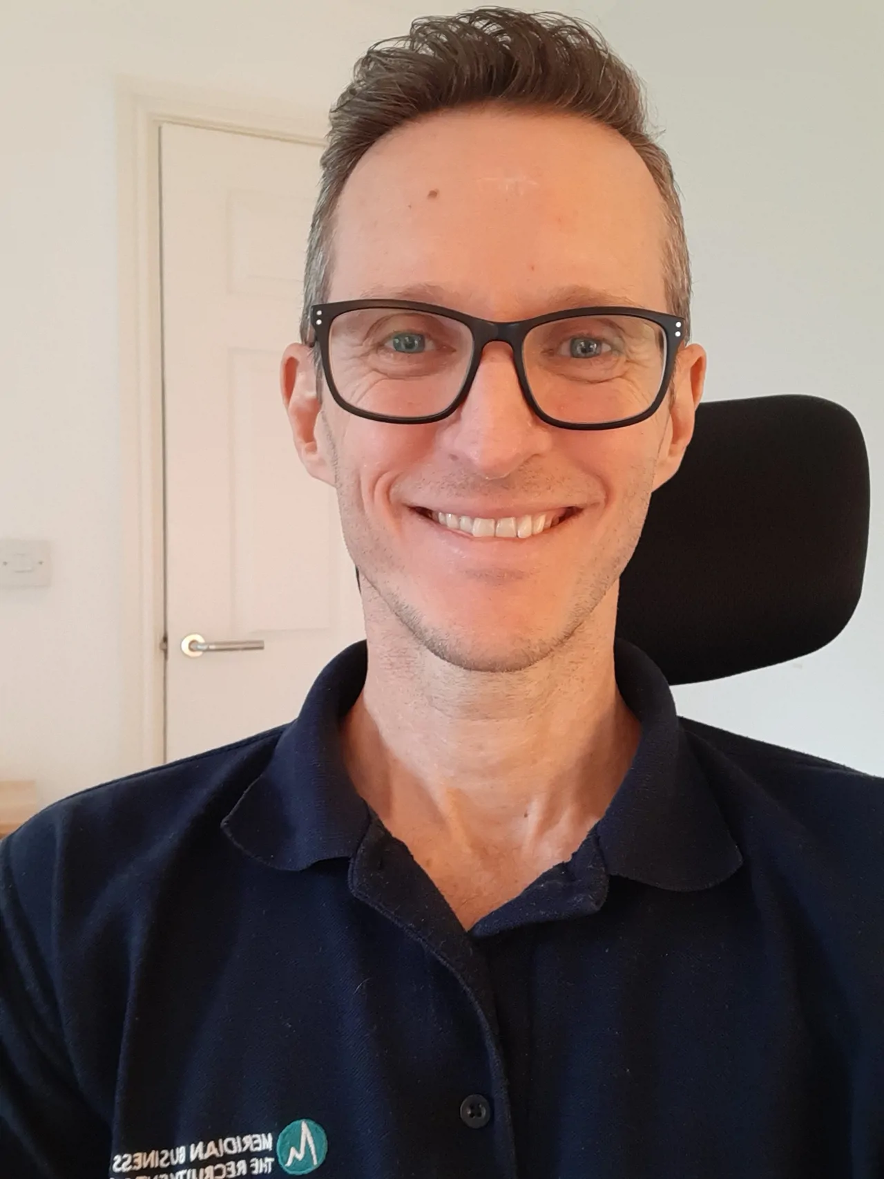 Man wearing glasses in Meridian branded polo smiling for photo in his office chair