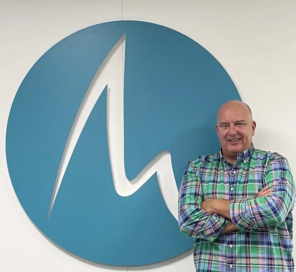 Man standing with arms crossed in front of a large blue Meridian Business Support logo on a white wall, wearing a green, blue, and white plaid shirt