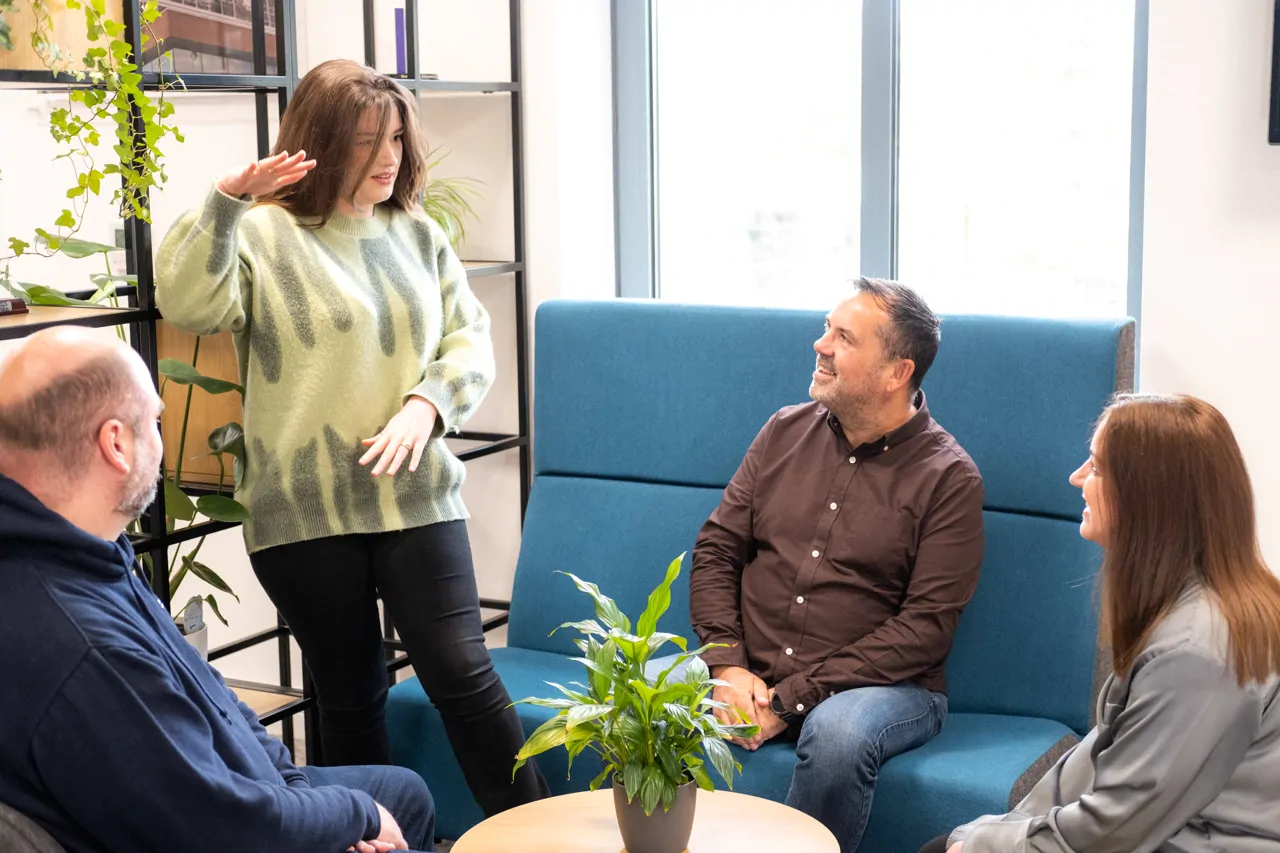 Three people sat on couches around a plant listening to woman stood up gesturing