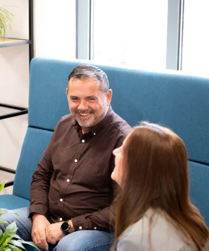 Two colleagues sat smiling together and talking on comfy blue seat at work