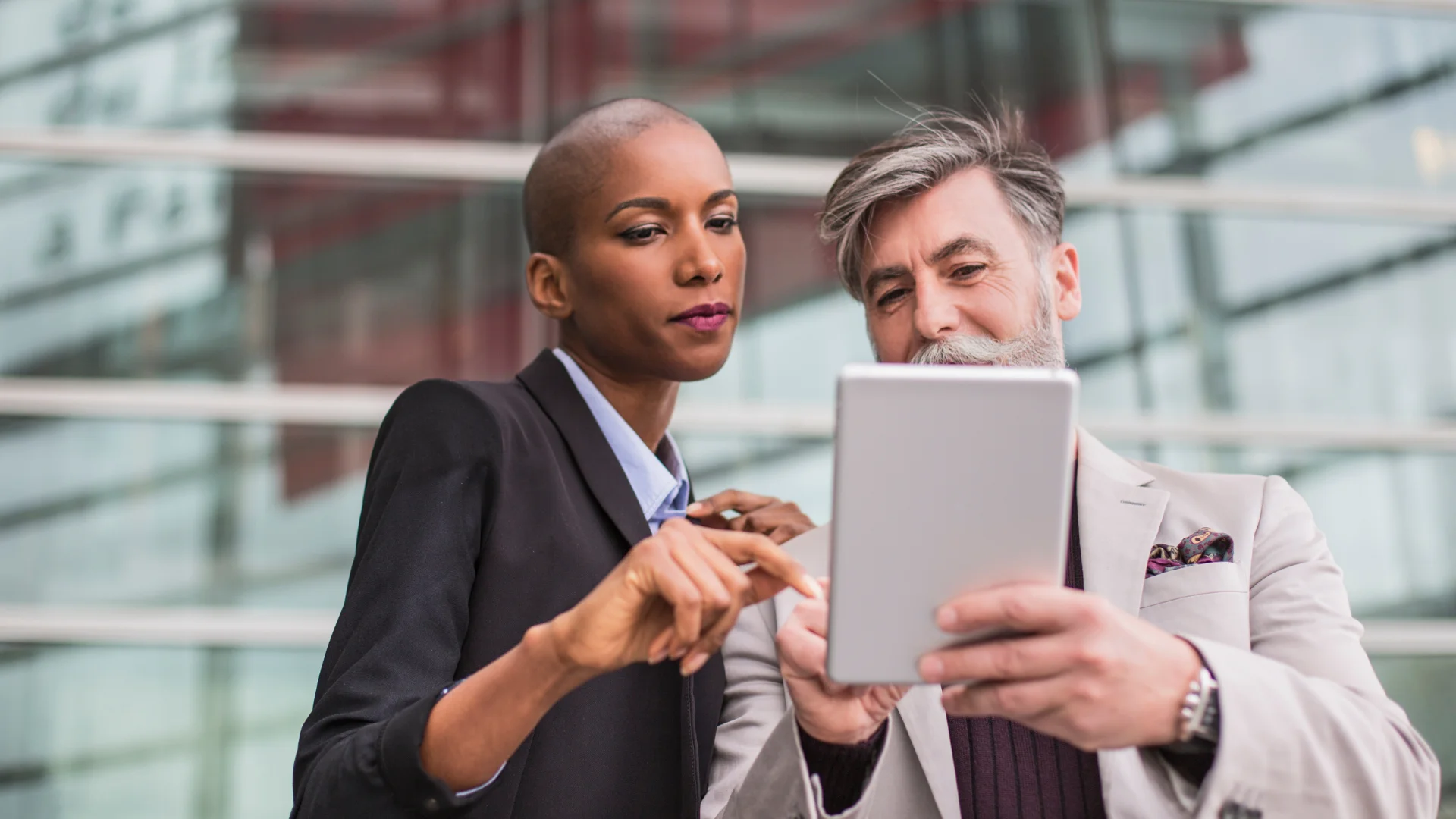 Two financial professionals reviewing reports and data on a tablet outside a corporate office