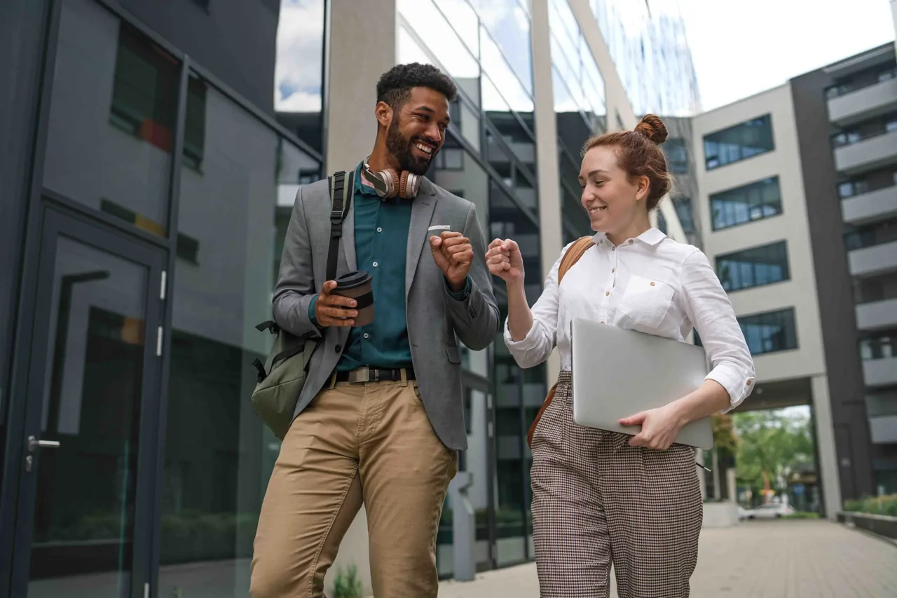 Two young professionals walking between office buildings. Both smiling and giving fist bumps. 