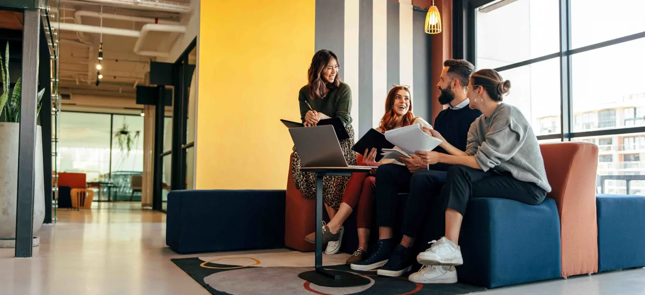 Four young office workers in a colourful office on a sofa laughing