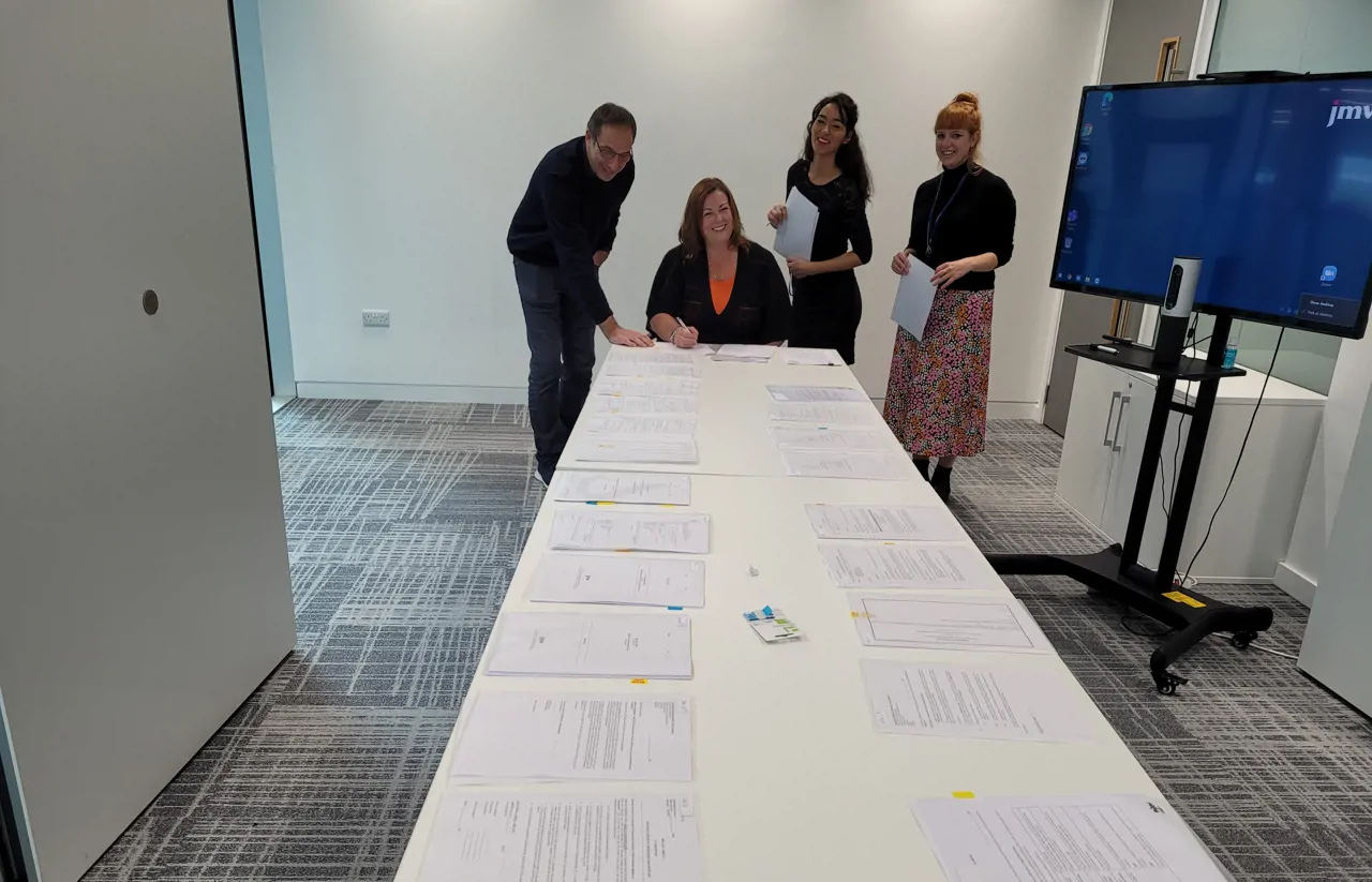 Contracts laid out on a table with Meridian director signing papers at the end of the table