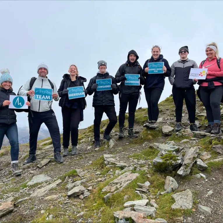 Group of people stood in a line on a mountain side holding up signs for a sponsored charity hike