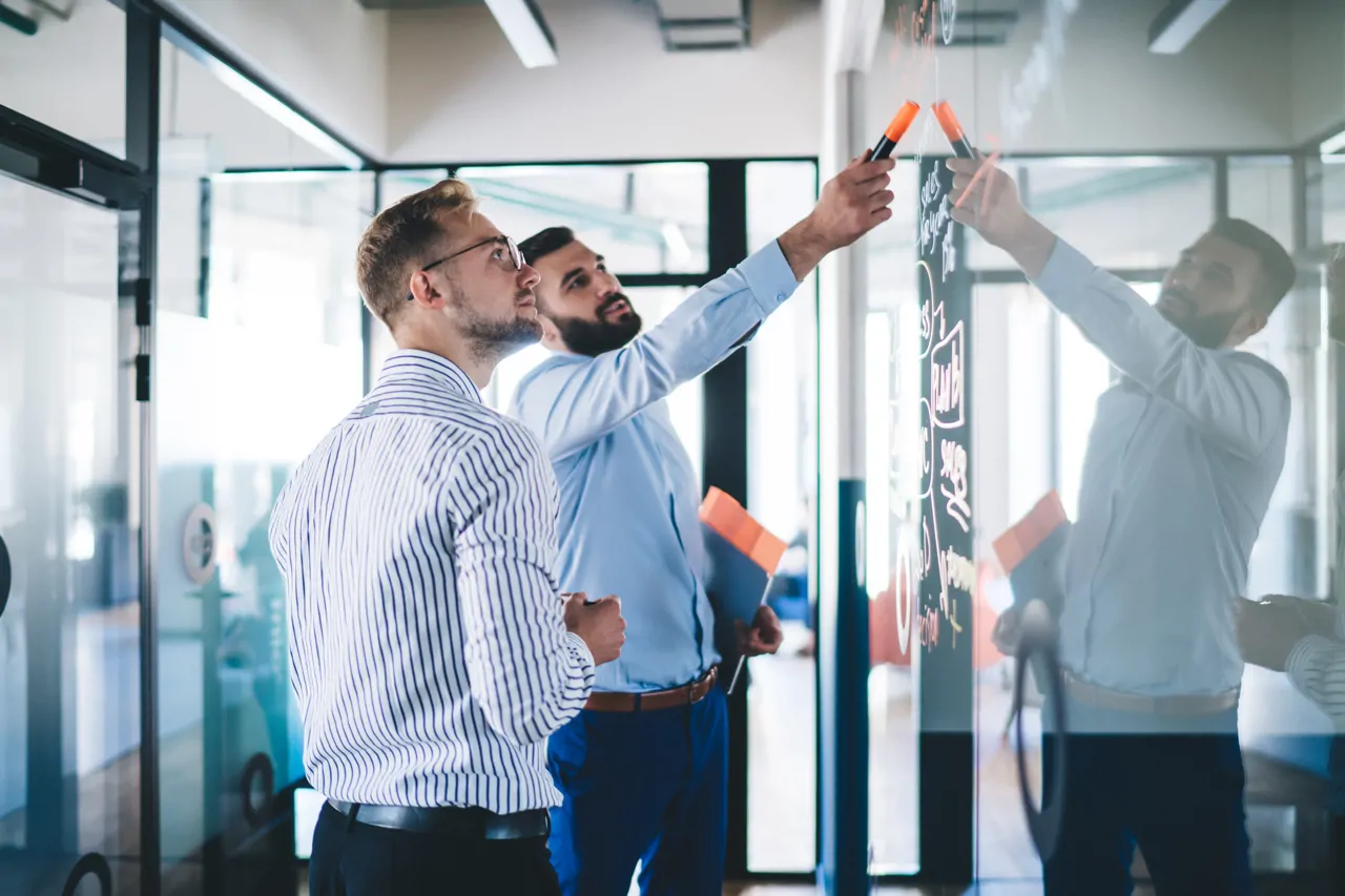 Confident male employee pointing on notes standing with colleague near glass creating infographic for conversation, skilled managers discussing sketches 