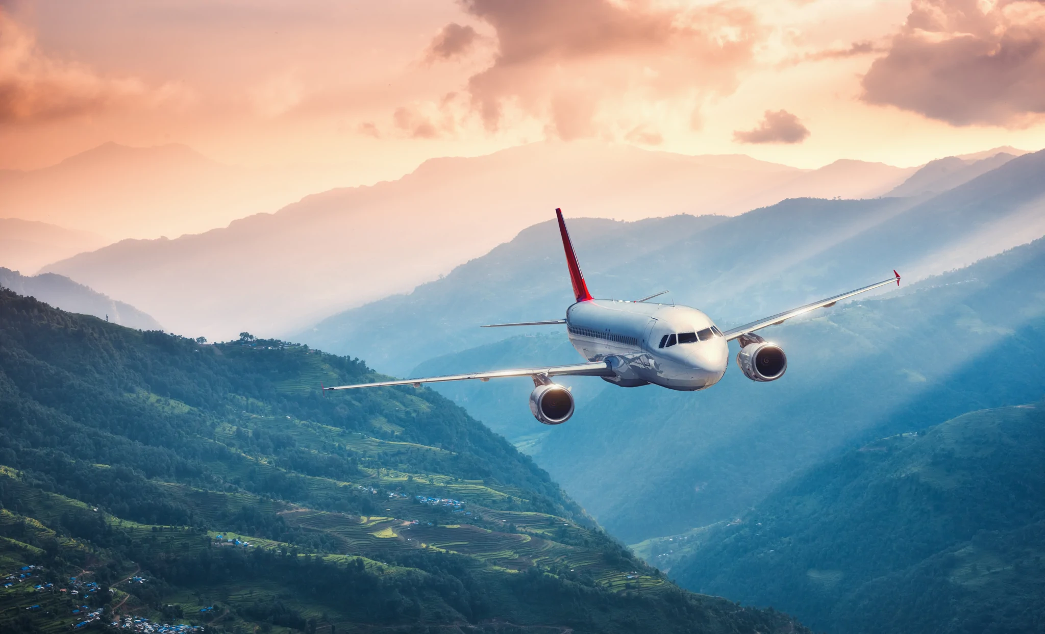 Aircraft is flying over green hills against mountains with yellow sunbeams at sunset. 