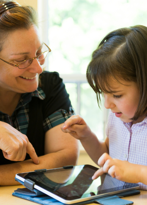 A young child playing on an ipad while a teacher looks on