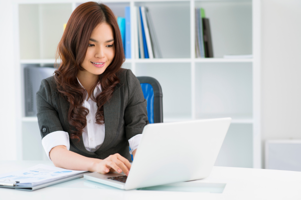 A lady wearing a white shirt and grey blazer sat at a desk looking at a laptop screen