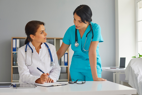 Two female nursing professionals speaking to each other as one writes on paper