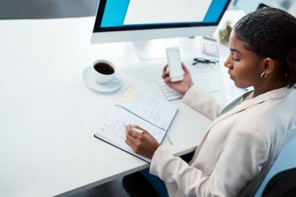 A lady sat at a desk writing in a notepad and holding a mobile in the other hand