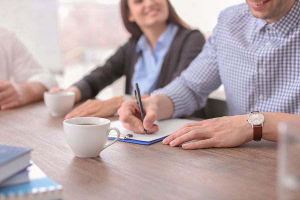 A man and lady sat around a desk whilst the man writes on a clipboard and paper