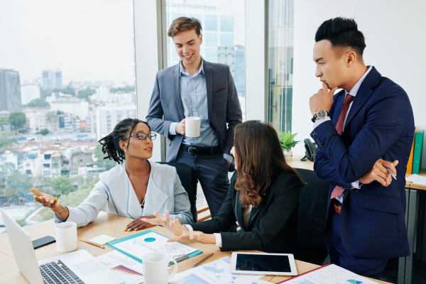 Two men stood up by two sitting women having a conversation in an office environment