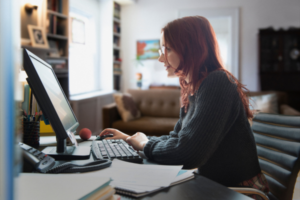 A lady sat at a desk wearing a black knitted jumper and glasses looking at her computer screen