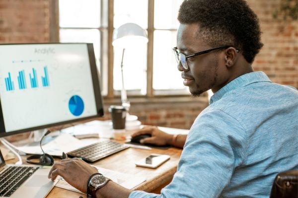 A man wearing glasses sat at a desk with a computer screen displaying a graph