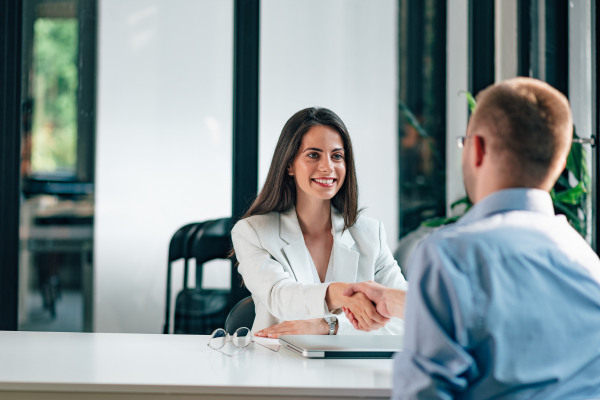 A lady wearing a white blazer shaking hands with a man sat opposite her