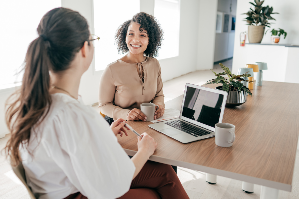 Two ladies sat at a desk with a laptop between them looking at each other