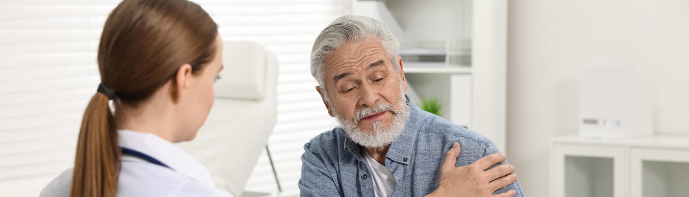 A female healthcare professional speaking to an elderly man who holds his shoulder