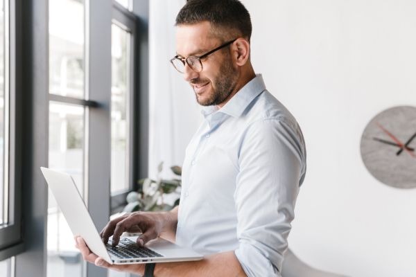 A man wearing glasses and a shirt holding a laptop and typing on it