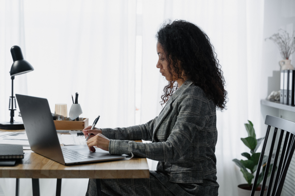 A lady sat at a desk with an open laptop next to her as she writes on paper