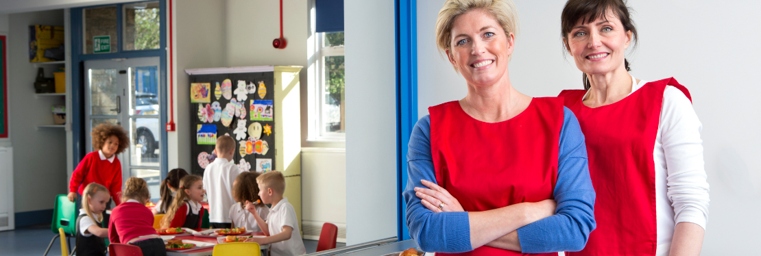 Two school cooks smiling while children sit in the background eating their dinner