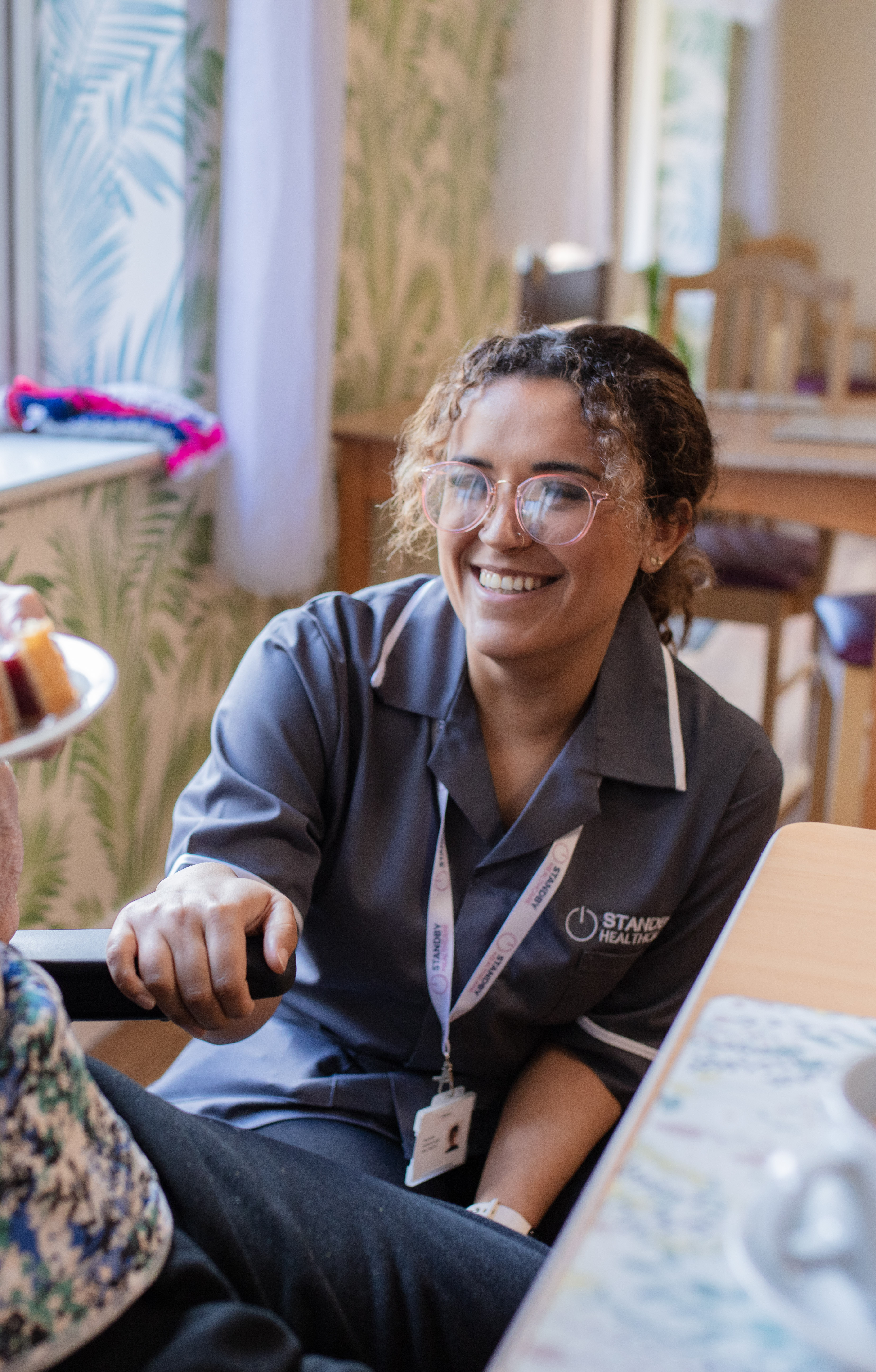 Carer leaning against table with hand on chair arm rest, smiling at person off camera.