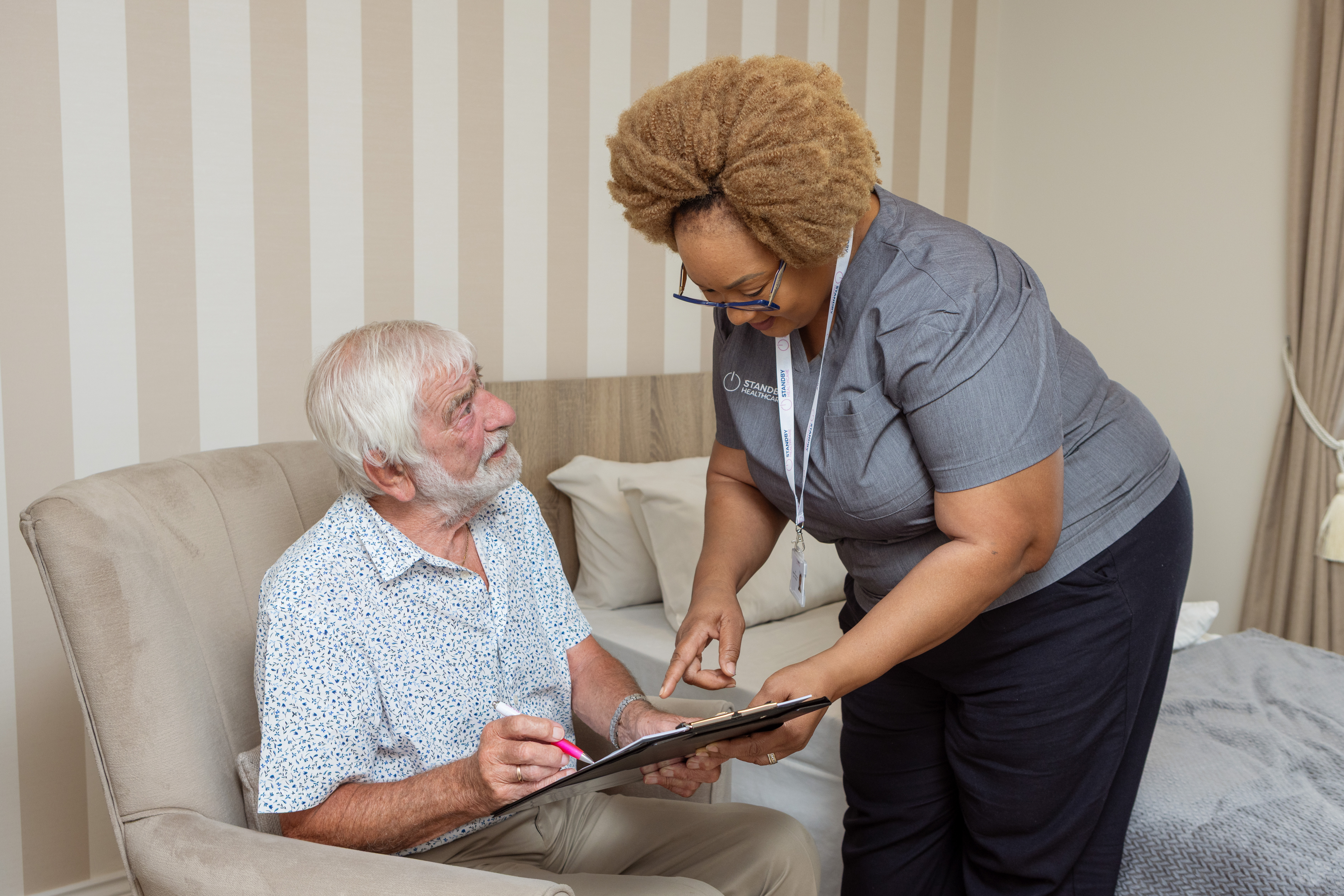 Woman in healthcare tunic leaning down, holding a clip board, smiling at an elderly man