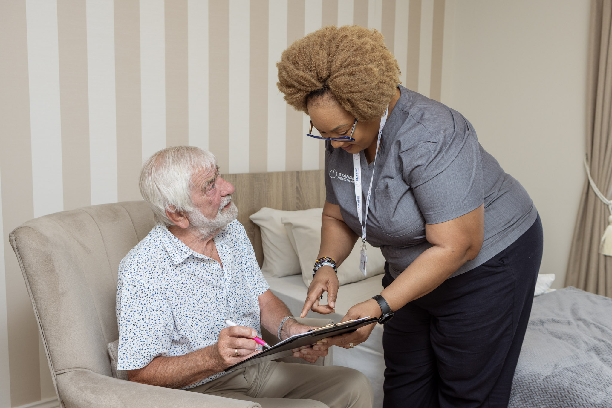 Woman in healthcare tunic leaning down, holding a clip board, smiling at an elderly man