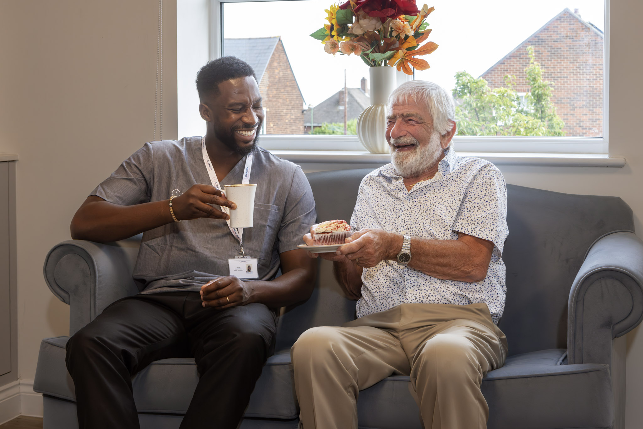 A male care staff member sat with an elderly man, smiling and sharing tea and cake