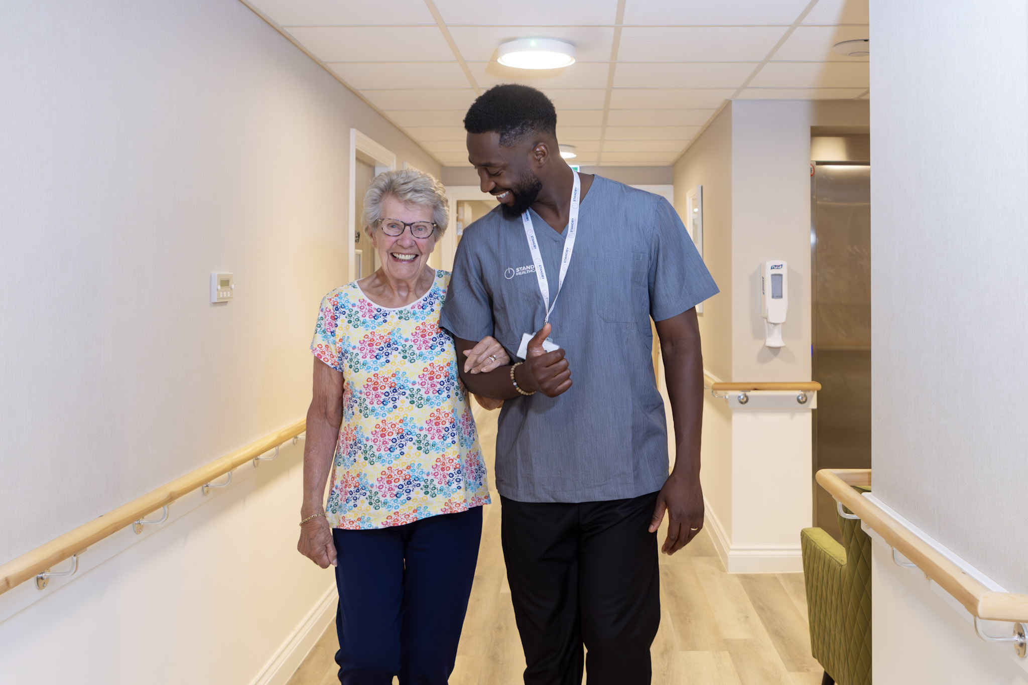An elderly woman, smiling, linking arms and walking with a male care staff worker 