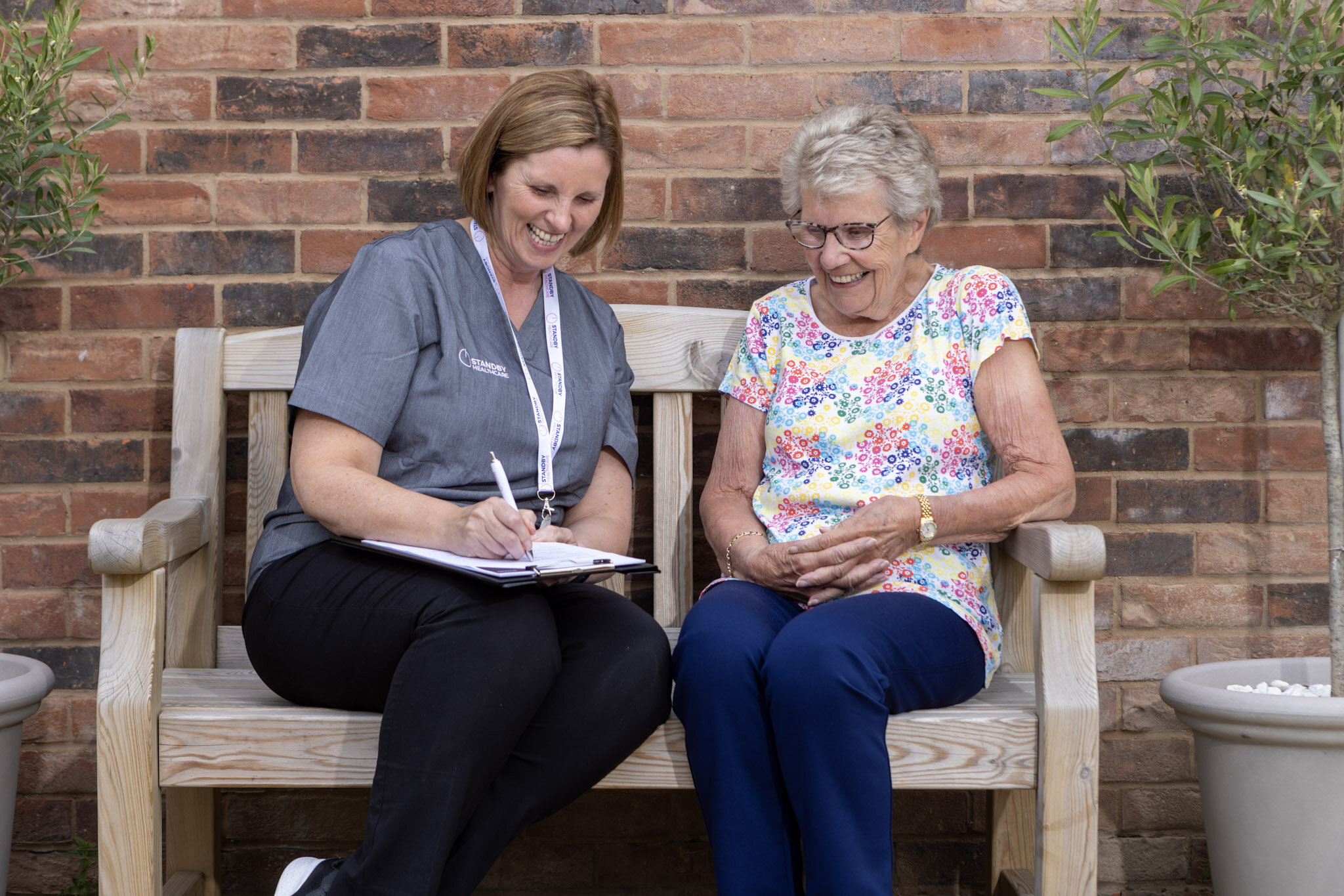 A female care staff member speaking to an elderly lady, both of whom are sat on a bench