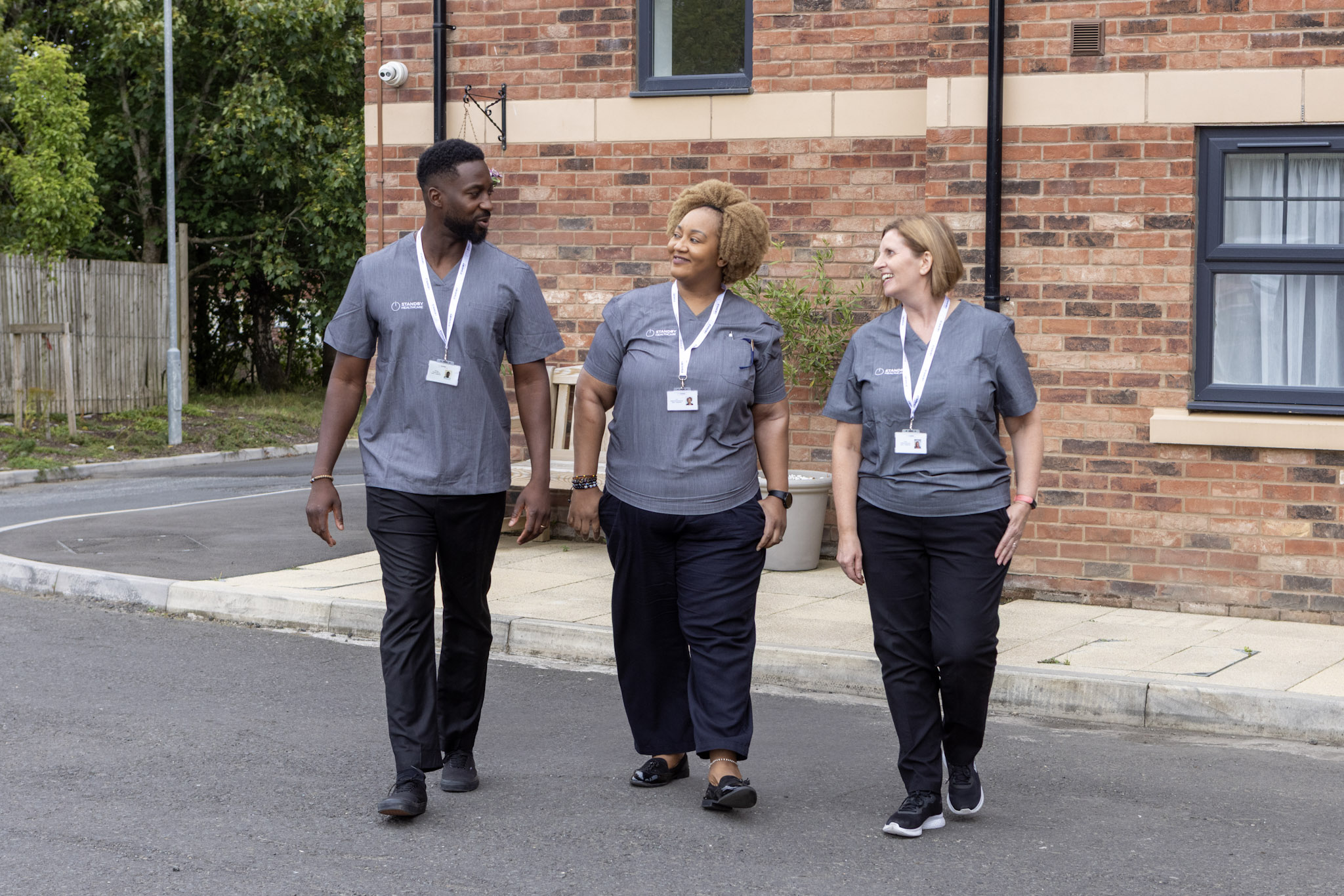One male and two female care staff, wearing grey tunics, walking outside together and smiling