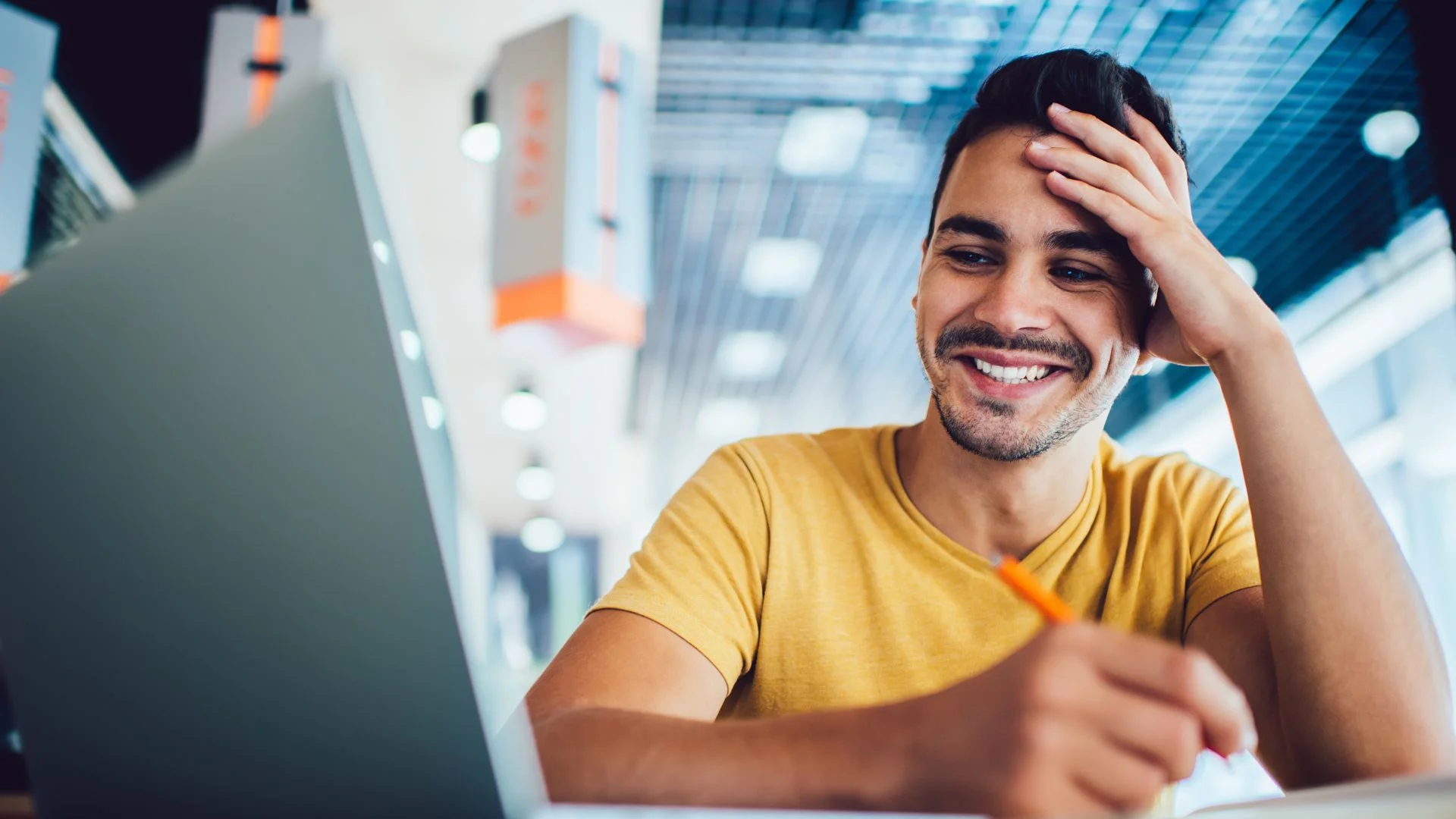 Smiling man in yellow shirt using laptop and writing notes at desk in modern office environment